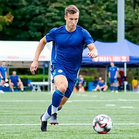 an athletic man playing soccer He should be shown running on a soccer field while kicking a soccer ball during a match